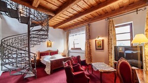 Una habitación con techo de madera, una escalera de caracol, una cama con tapicería roja, una mesa con mantel de encaje y una silla.