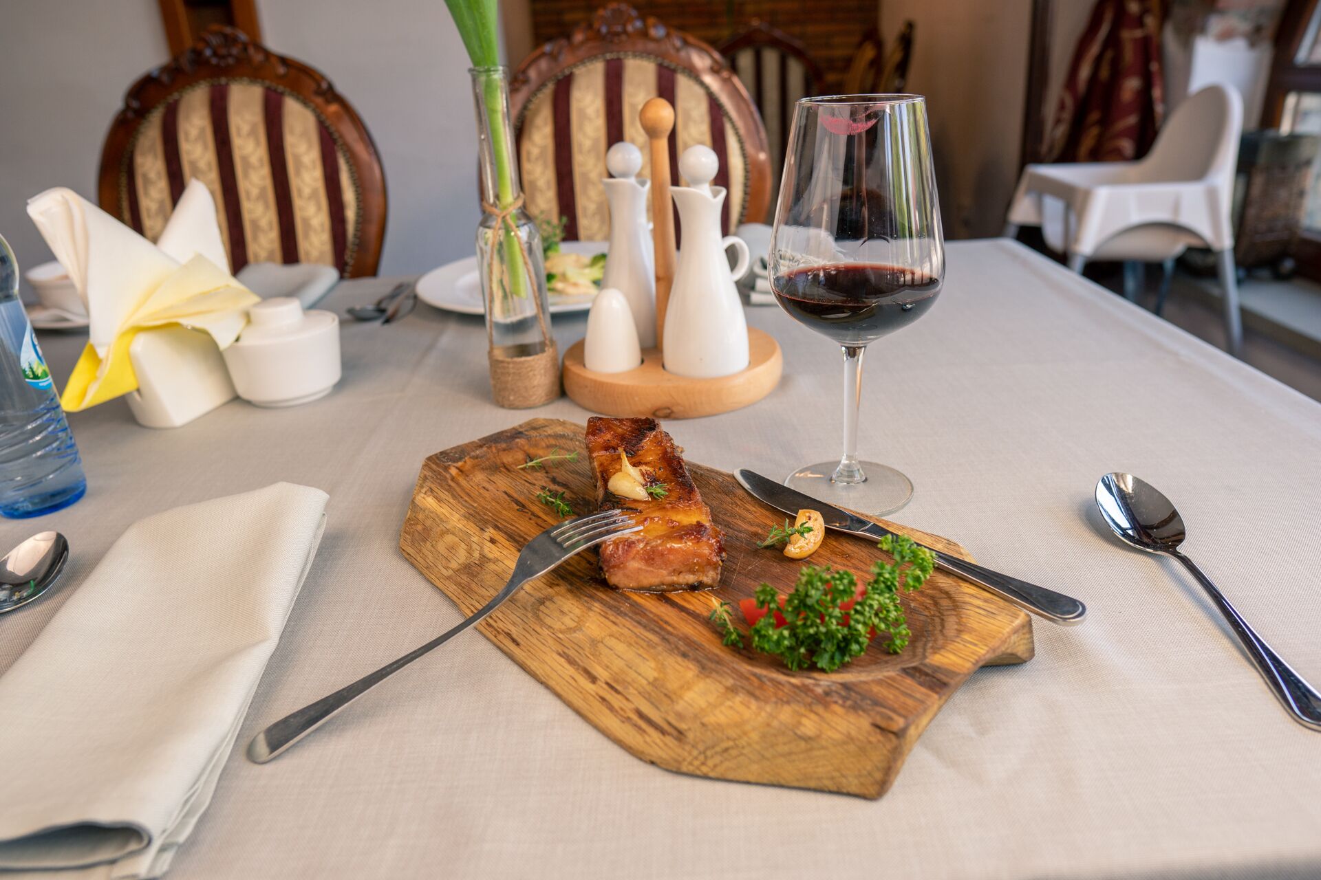 Una mesa de comedor con una tabla de madera que sostiene un trozo de carne, una copa de vino tinto y un tenedor. Hay saleros y pimenteros, una botella de agua y una servilleta sobre la mesa.