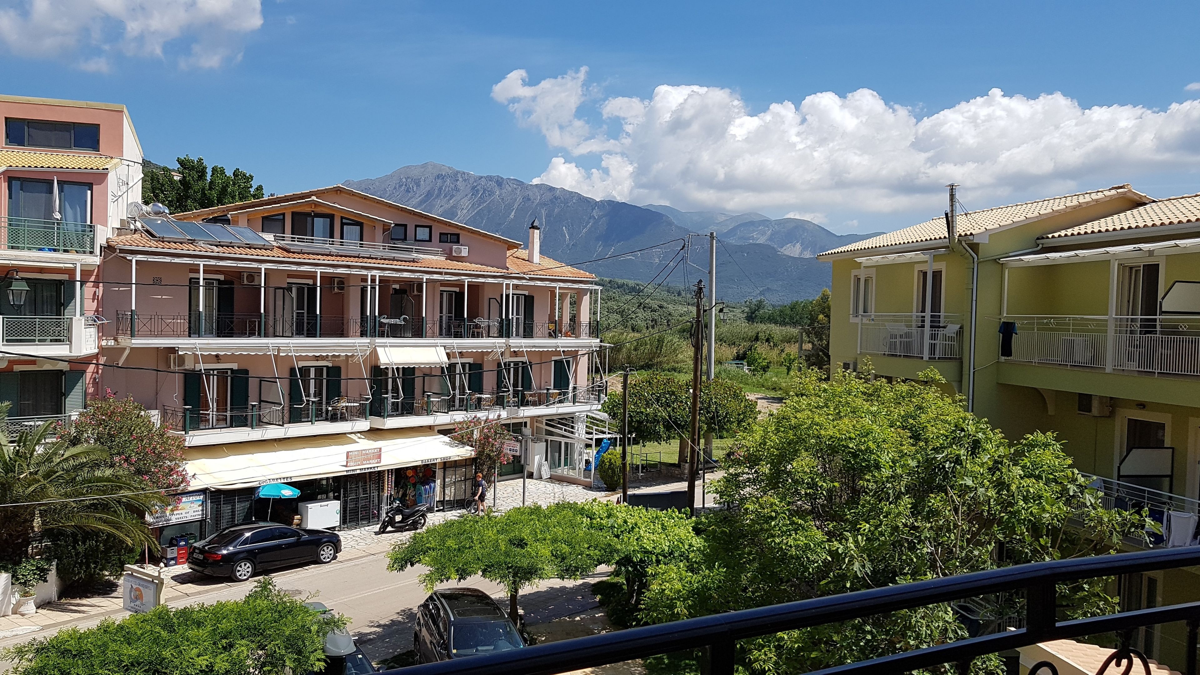 A multi-story building with balconies, a visible ground floor with a sign for a bakery shop, and a mountainous backdrop.
