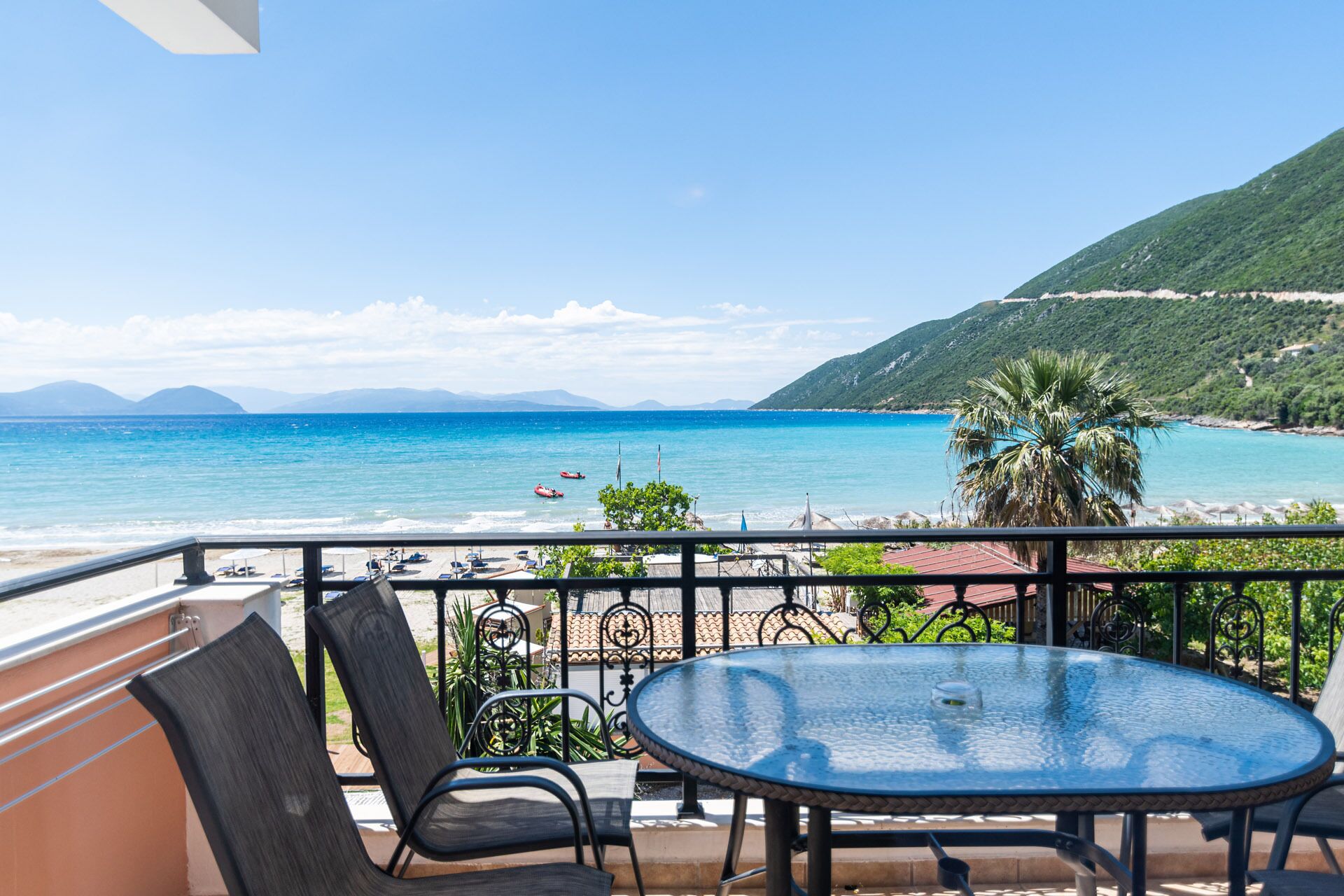 A balcony with a glass table and chairs overlooking a beach with clear blue water and distant mountains.