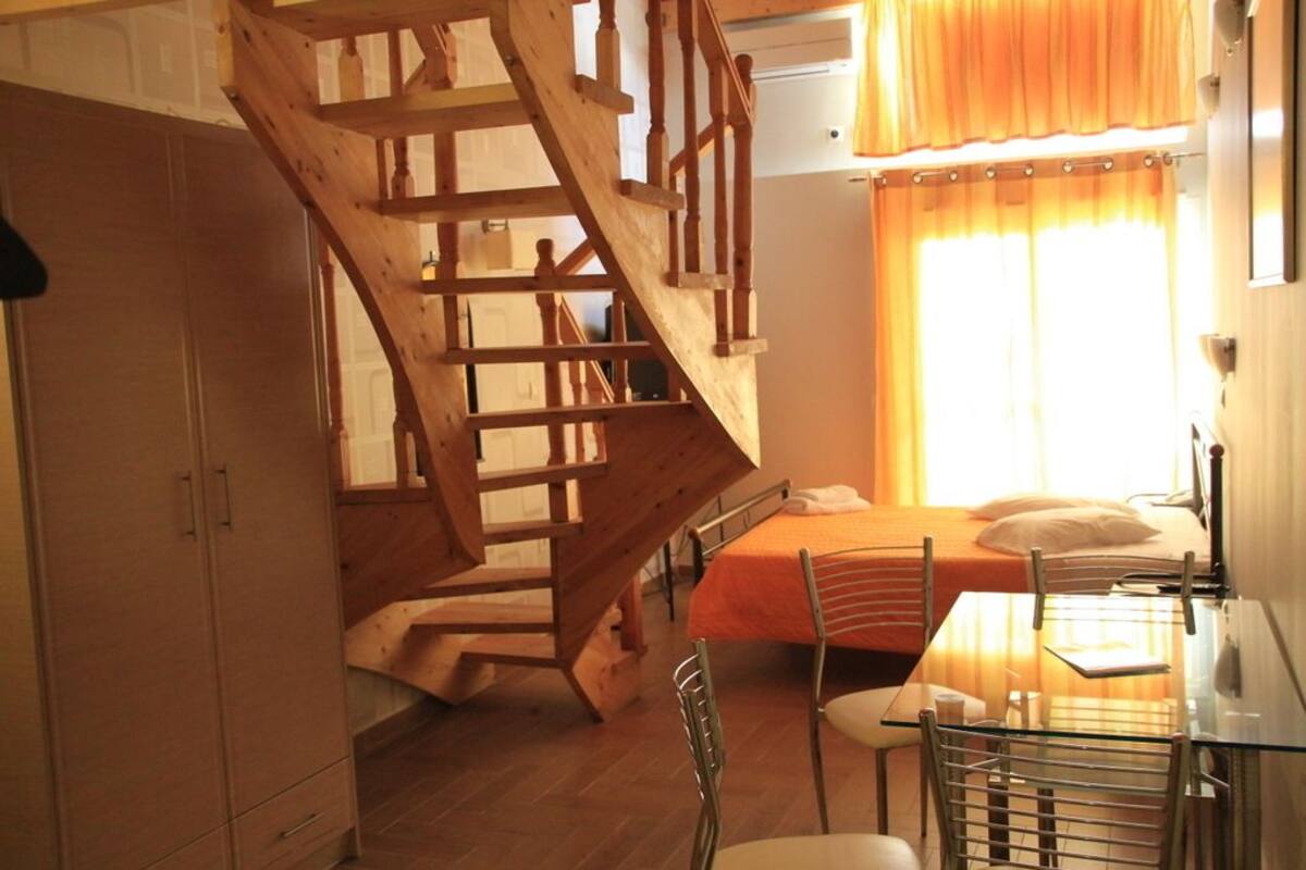 Un dormitorio con escalera de madera, cama con colcha naranja, una mesa de vidrio y una silla.