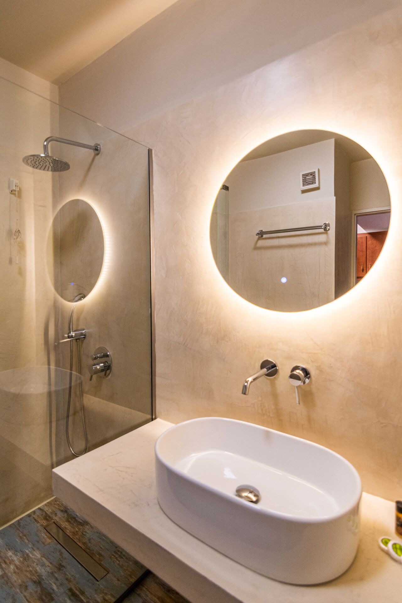 A modern bathroom with a glass-enclosed shower, a round mirror with a light, and a white sink.