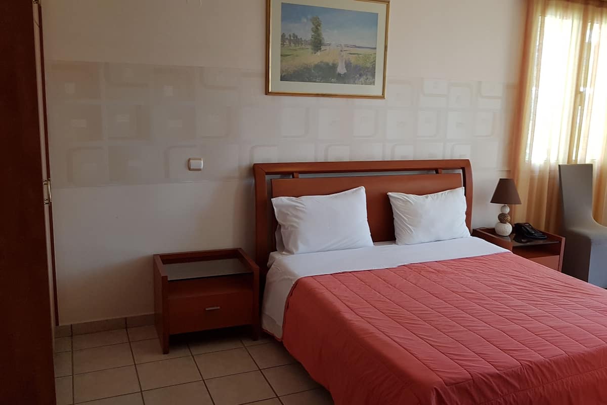 Habitación de hotel con cama de madera, almohadas blancas, colcha roja, mesita de noche con lámpara, una silla y un cuadro en la pared.