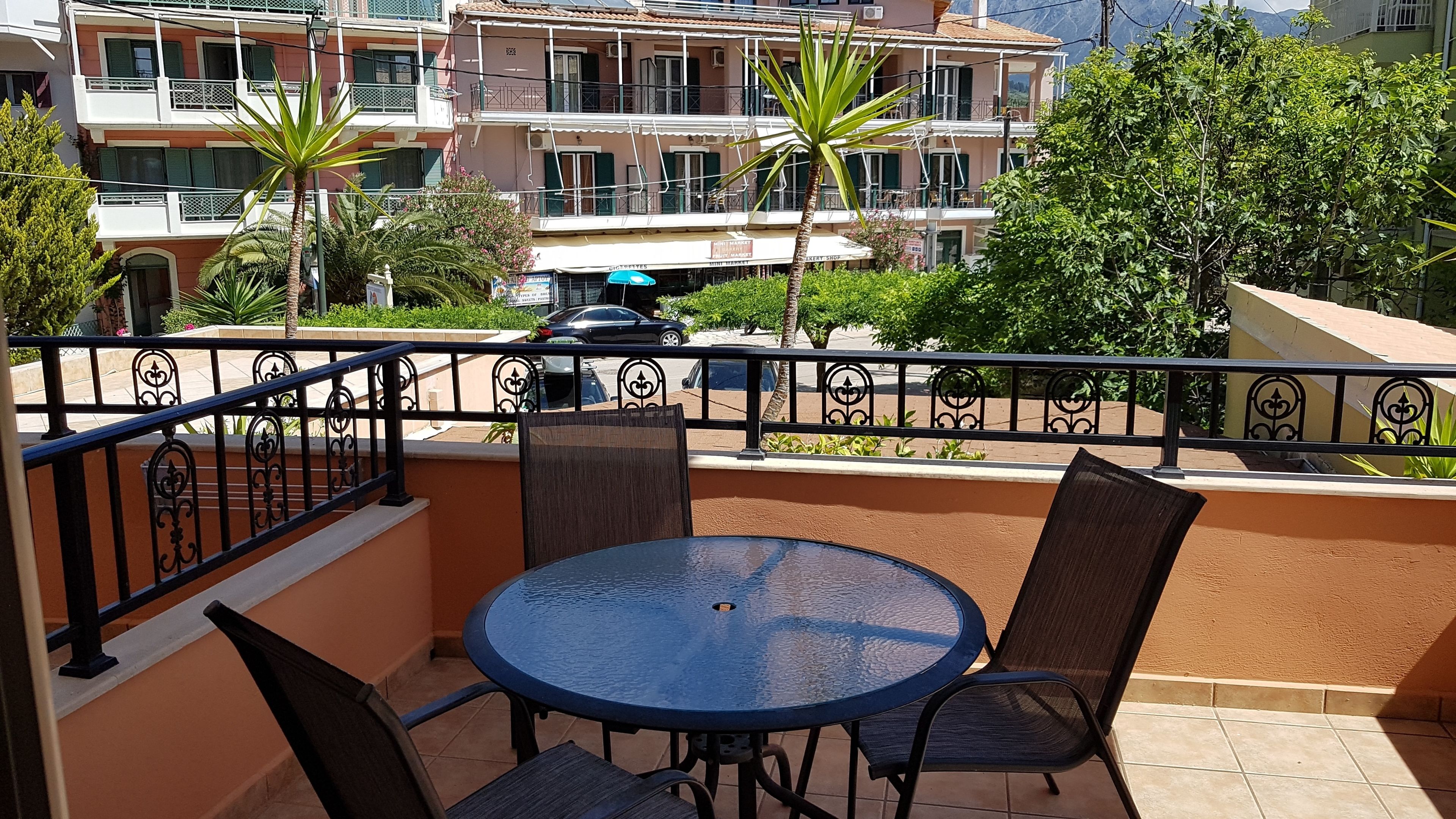 Un balcon avec une table ronde et des chaises, donnant sur une rue bordée de voitures garées et d’immeubles.