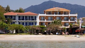 Un hotel frente a la playa con varios balcones, una playa de arena y montañas al fondo.