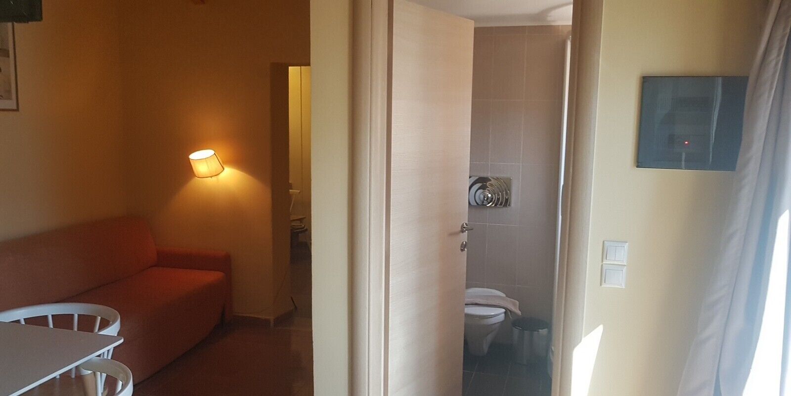 A hotel room with a sofa, a table, a lit lamp, a toilet, and a shower area.