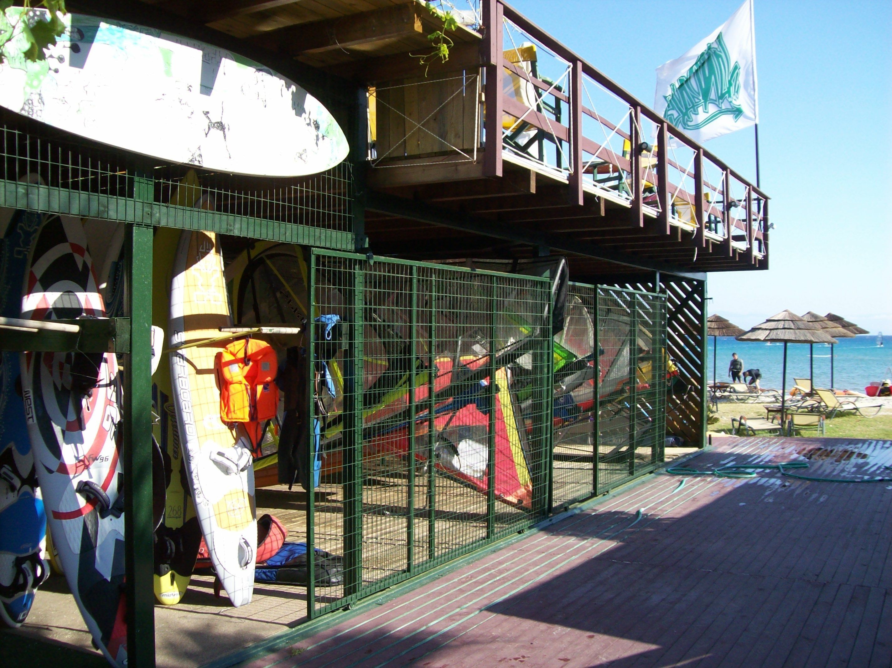 A beachside building with a covered deck area, a fenced enclosure with surfboards, and a view of the beach and ocean.