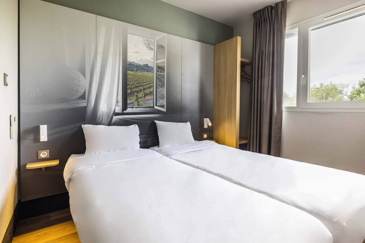 Ein Doppelbett mit weißen Laken in einem Hotelzimmer mit großem Fenster, einem Holfnachtkästchen und Blick auf einen Weinberg.