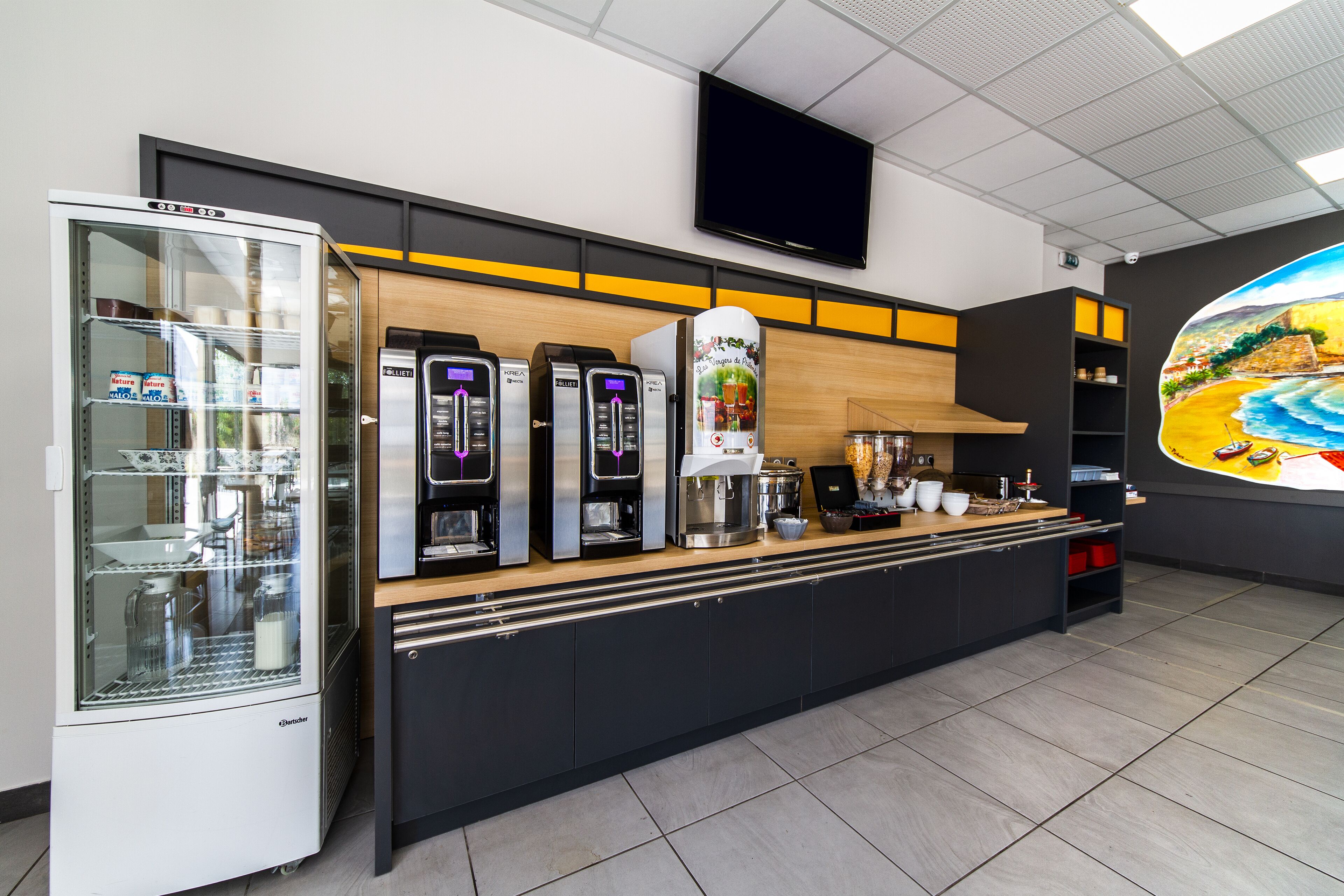 A modern kitchen area with a coffee machine, a refrigerator, and a flat-screen TV mounted on the wall.