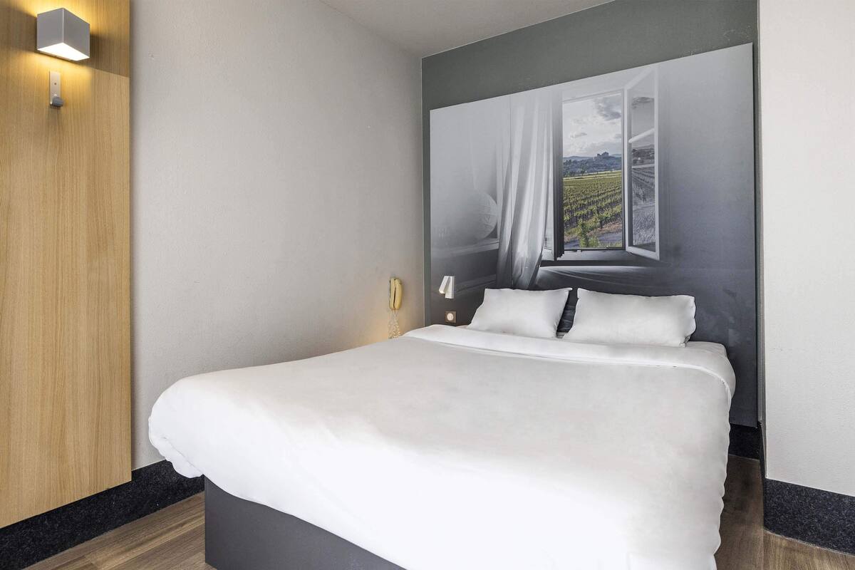 Ein ordentlich bezogenes Bett mit weißen Leinen in einem modernen Zimmer mit Holzvertäfelung und einem großen Fenster mit Blick auf einen Weinberg.