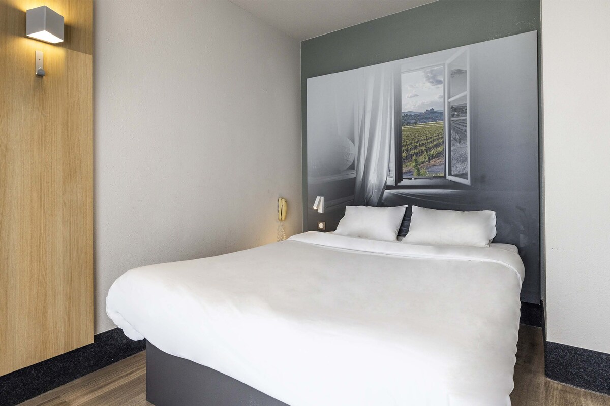 A neatly made bed with white linens in a modern room with a wooden panel and a large window with a view of a vineyard.