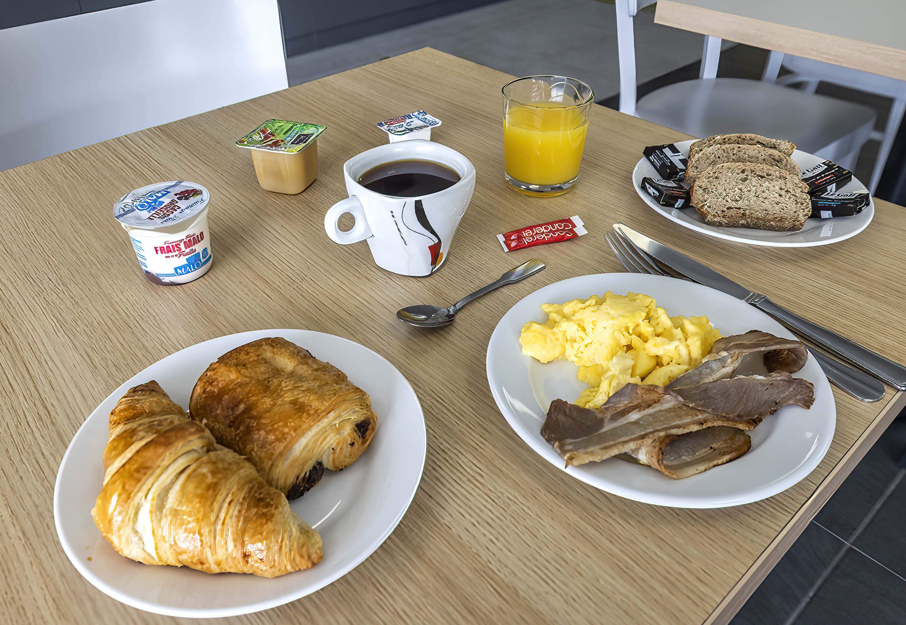 A breakfast table with croissants, scrambled eggs, bacon, a cup of coffee, a glass of orange juice, and a plate of bread.