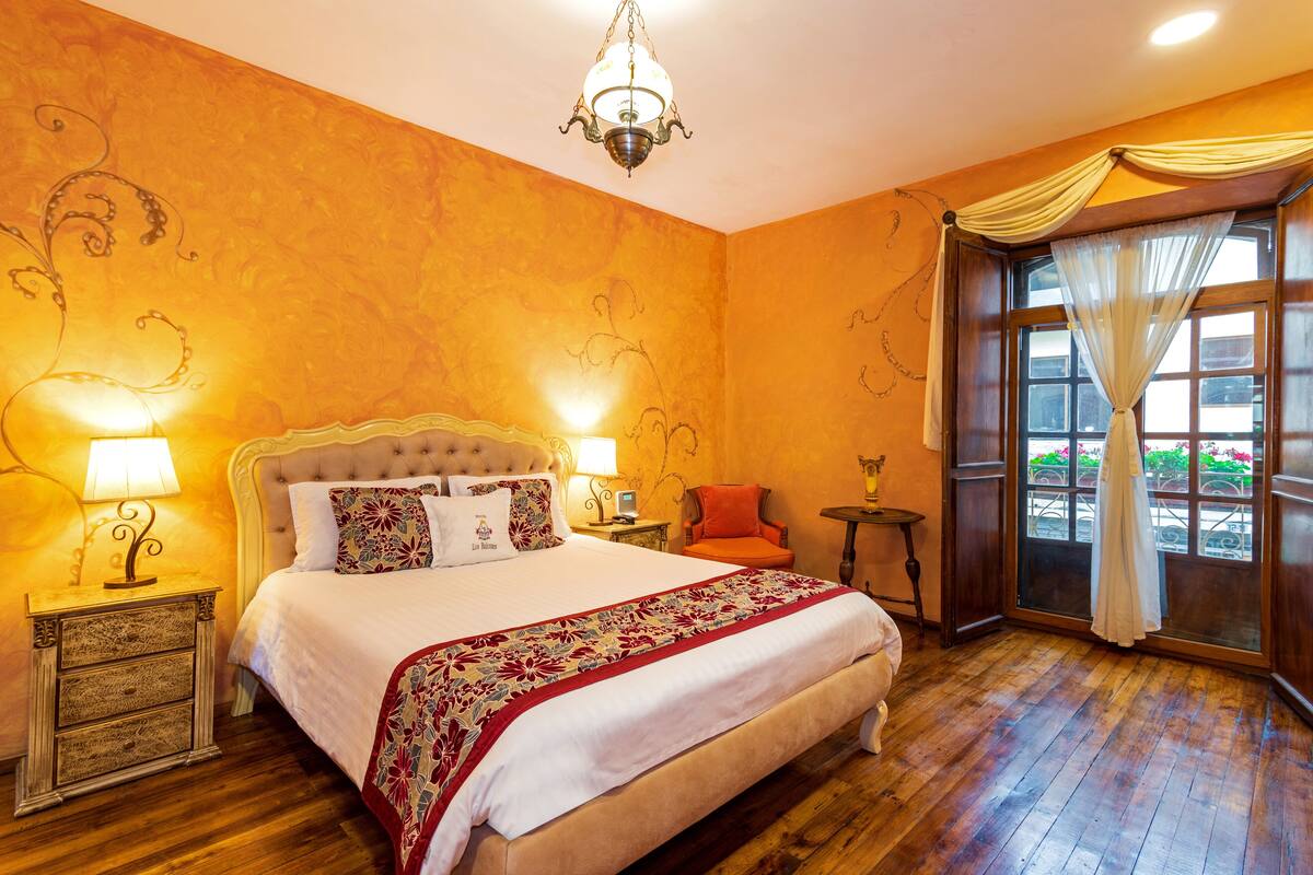 Una habitación de hotel con una cama grande, dos mesitas de noche con lámparas, una mesita pequeña, una silla y una ventana con cortinas.