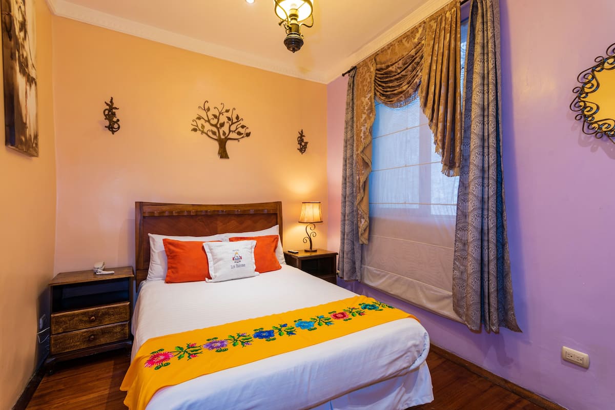 Una cama bien hecha con una colcha estampada con motivos florales, una mesita de noche con una lámpara y un adorno de árbol colgado en la pared.