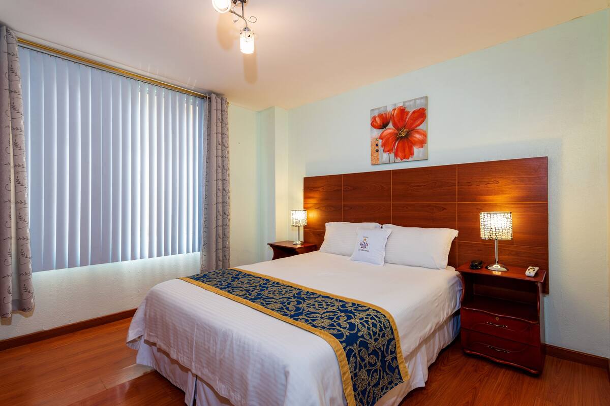 Habitación de hotel con una cama grande, cabecera de madera, dos mesitas de noche con lámparas, una ventana con cortinas y un cuadro con motivos florales en la pared.