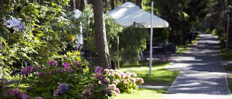 A pathway lined with trees and flowering plants, leading to a covered area with tables and chairs.