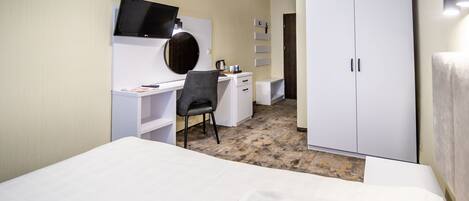 Habitación de hotel con cama, escritorio con silla, televisor, armario y una mesita con hervidor.