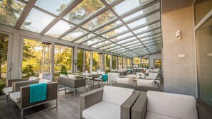 A spacious outdoor lounge area with wicker furniture and a glass ceiling, offering views of the surrounding trees. - Hotel Jantar (Ustka)