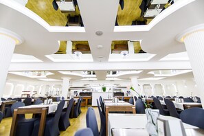A spacious dining area with multiple tables and chairs, a central service counter, and a high ceiling with recessed lighting. - Hotel Jantar (Ustka)