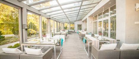 A covered outdoor dining area with glass walls, wicker furniture, and a view of the surrounding trees.