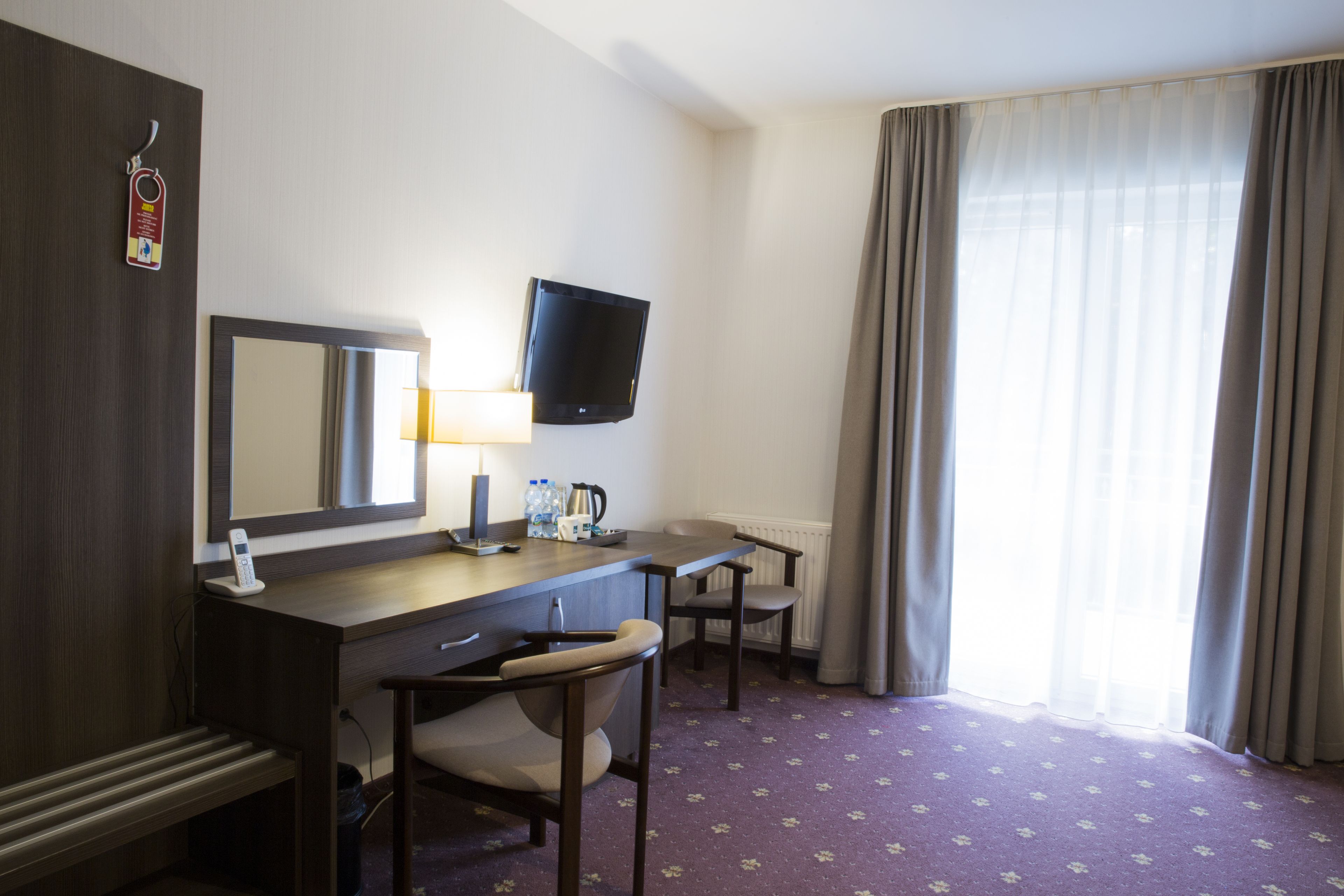 A hotel room with a desk, chair, television, and a large window with curtains.