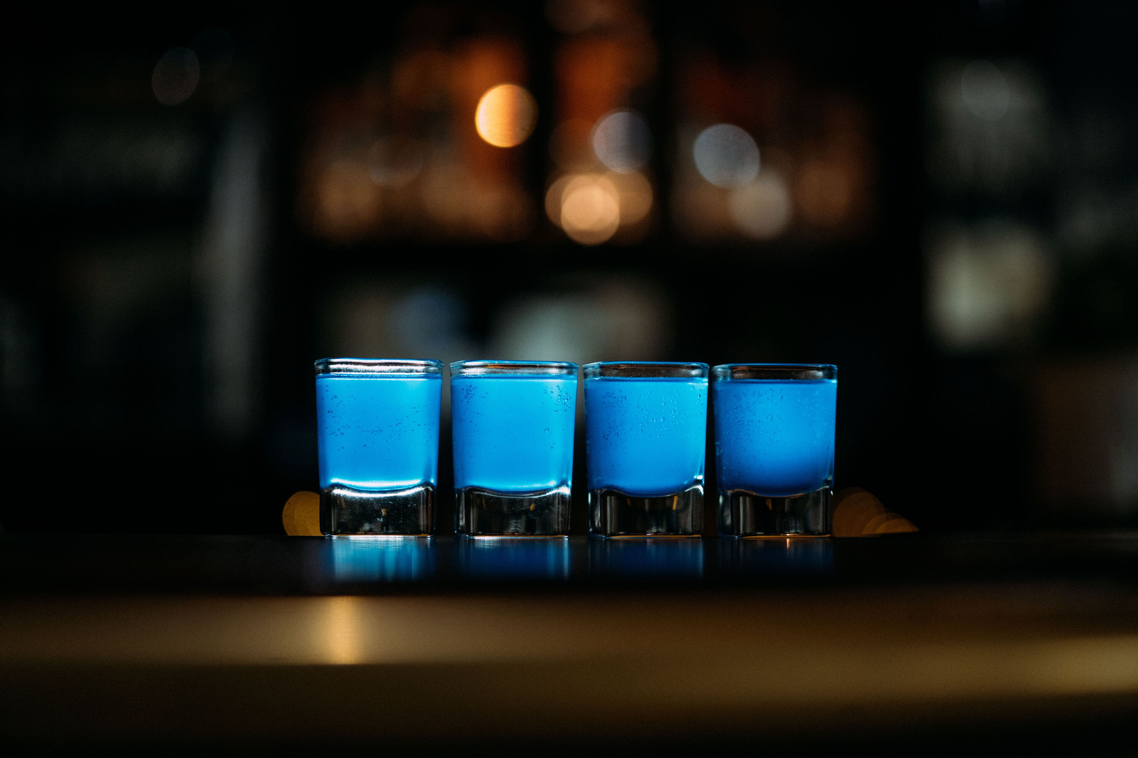 Quatre petits verres à shooter remplis d’un liquide bleu, posés sur une surface réfléchissante.
