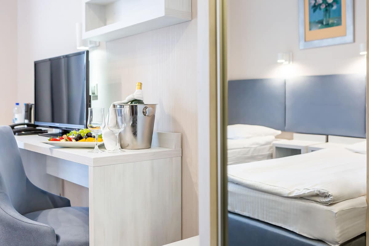 Une chambre d’hôtel comprenant un lit, un bureau avec un ordinateur, une chaise, un minibar et un coin repas avec une assiette de fruits et une bouteille de champagne.