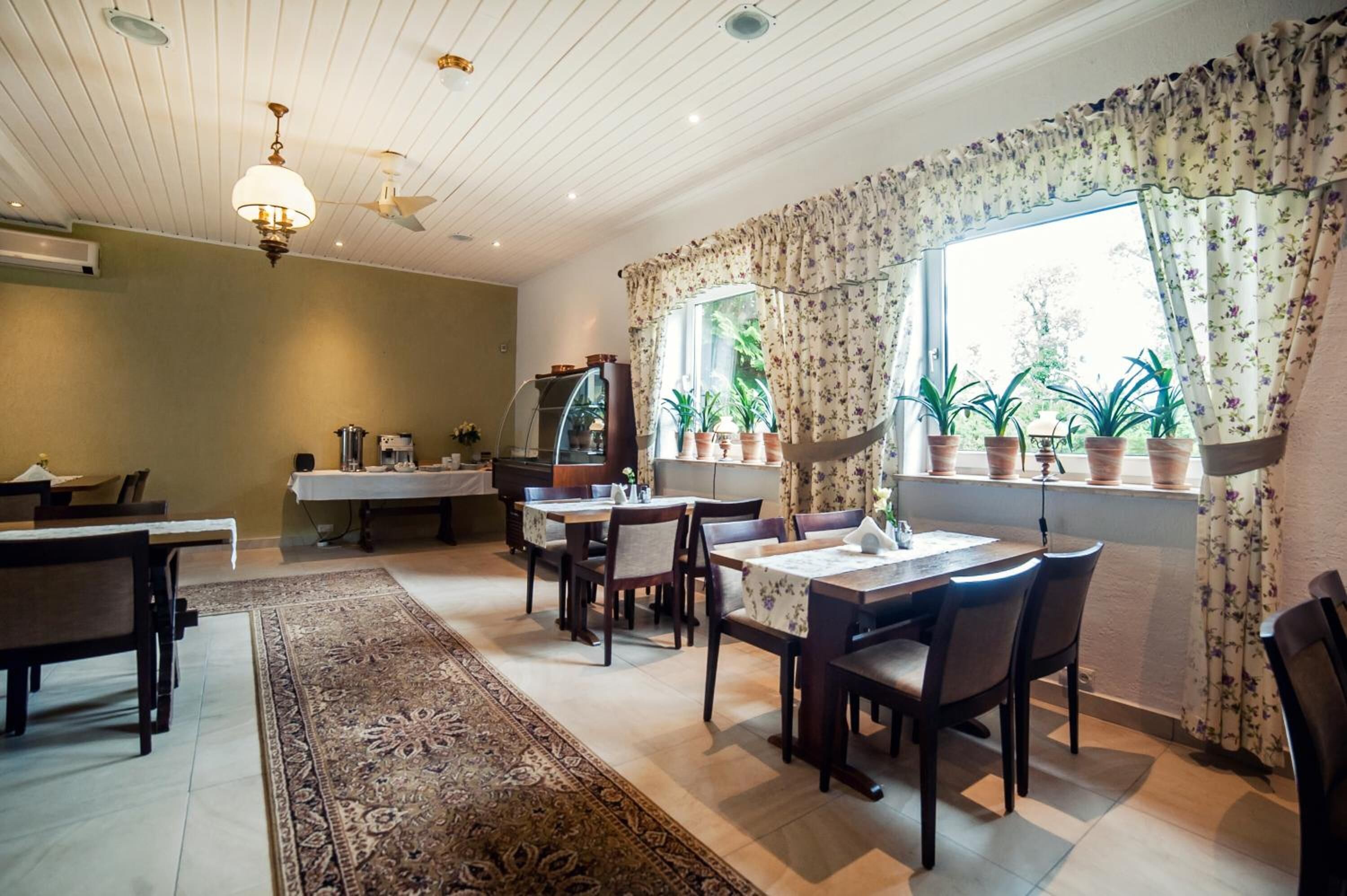 Uma área de jantar com mesas e cadeiras, um tapete grande, um ventilador de teto e uma janela com cortinas e plantas em vasos.