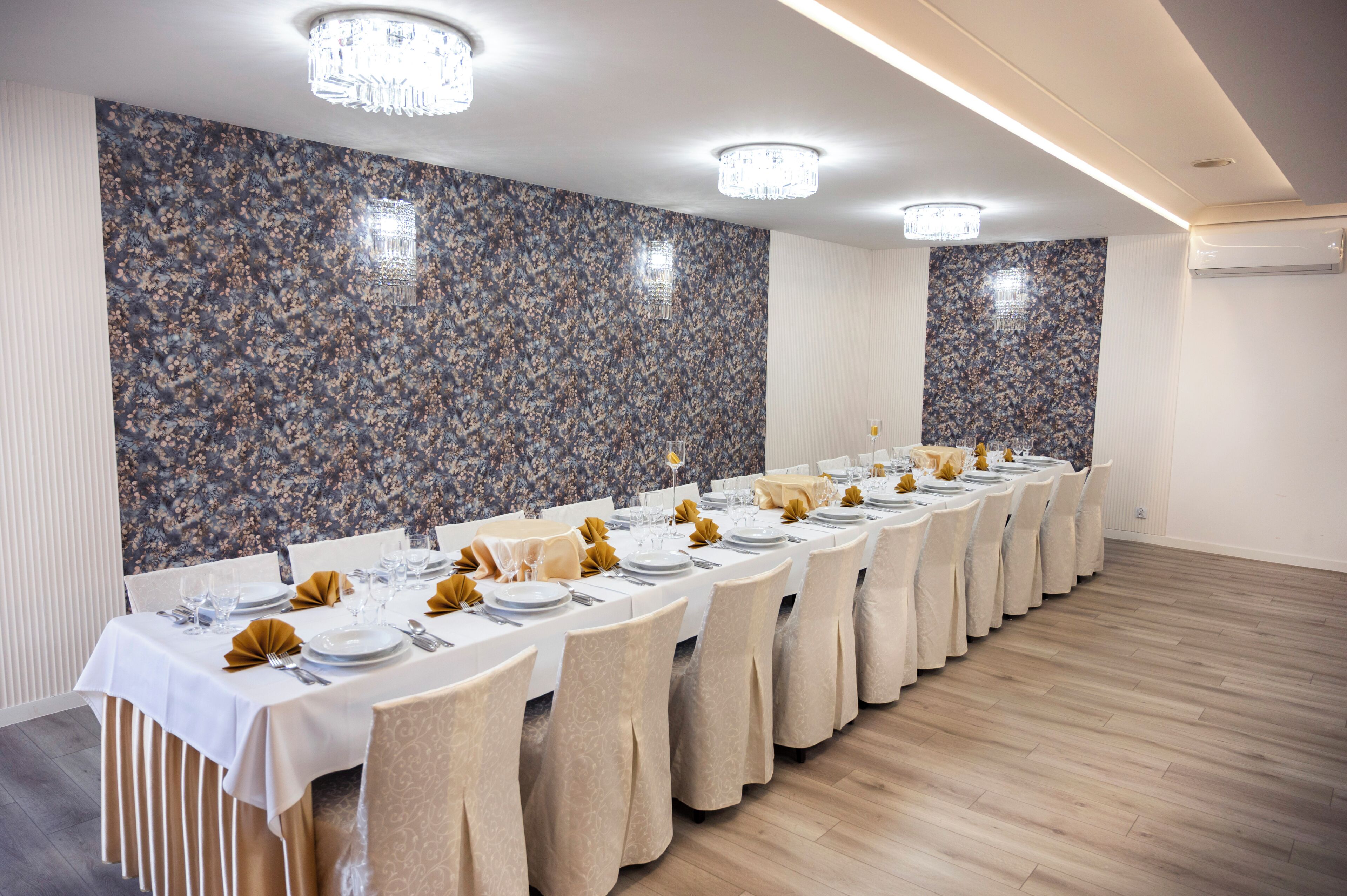 Una larga mesa de comedor con sillas, preparada para una comida, en una habitación con paredes y luces de techo decoradas.