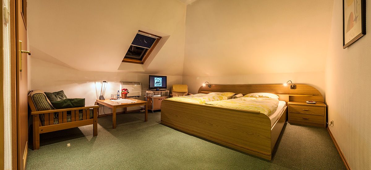 Une chambre avec un lit simple, un bureau avec une chaise, une petite table, une table de chevet, une télévision et une fenêtre donnant sur le ciel.