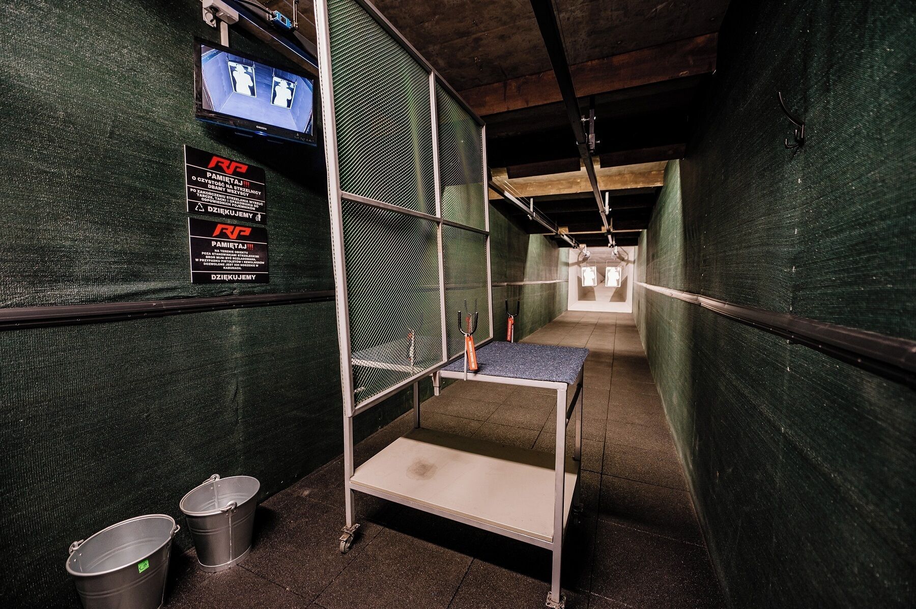 A narrow corridor with bunk beds, a metal table, and two buckets.