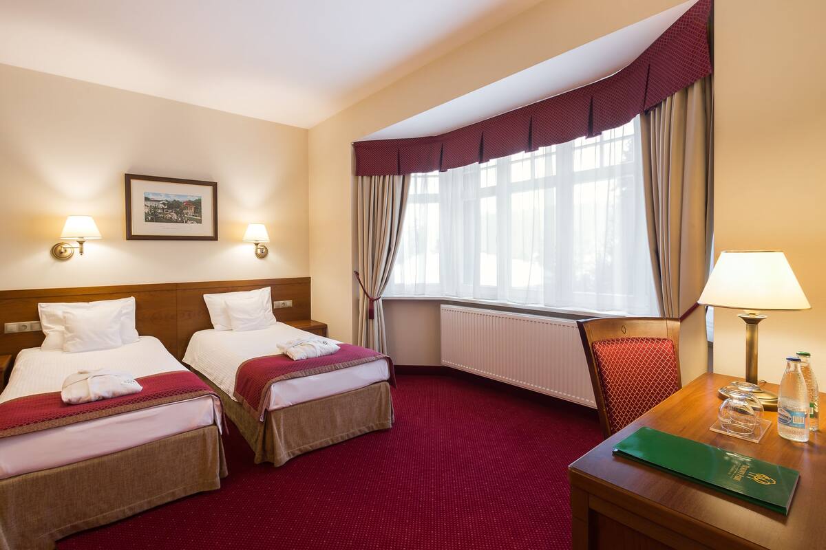 Une chambre d’hôtel avec deux lits, une tête de lit en bois, une moquette rouge, une fenêtre avec des rideaux, un tableau encadré au mur et une table avec une lampe et un dossier vert.