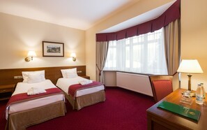 A hotel room with two beds, a wooden headboard, a red carpet, a window with curtains, a framed picture on the wall, and a table with a lamp and a green folder. - Bukowy Park Hotel Medical Spa (Polanica-Zdroj)