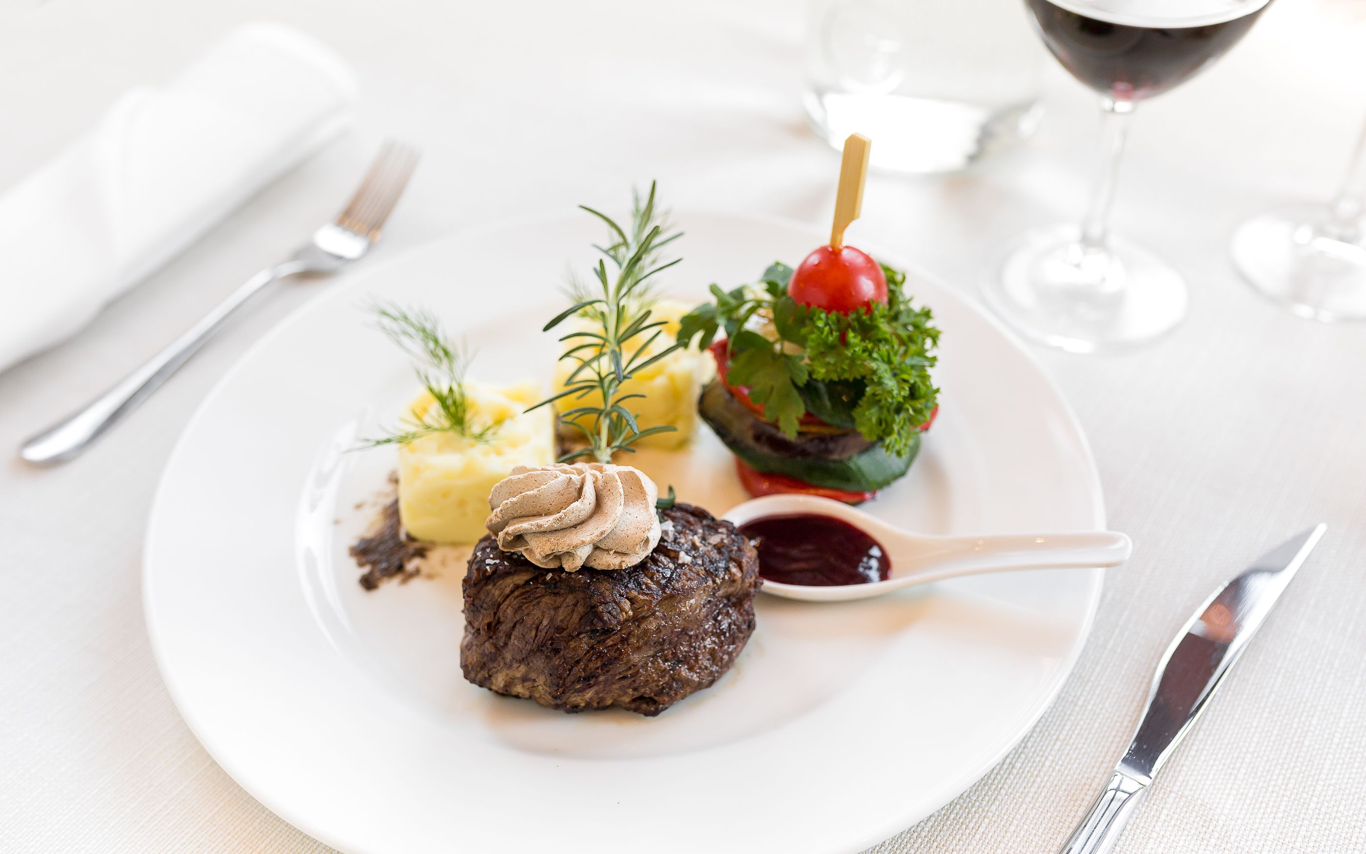 A plate with a steak, mashed potatoes, and a cherry tomato on a stick, accompanied by a glass of red wine.
