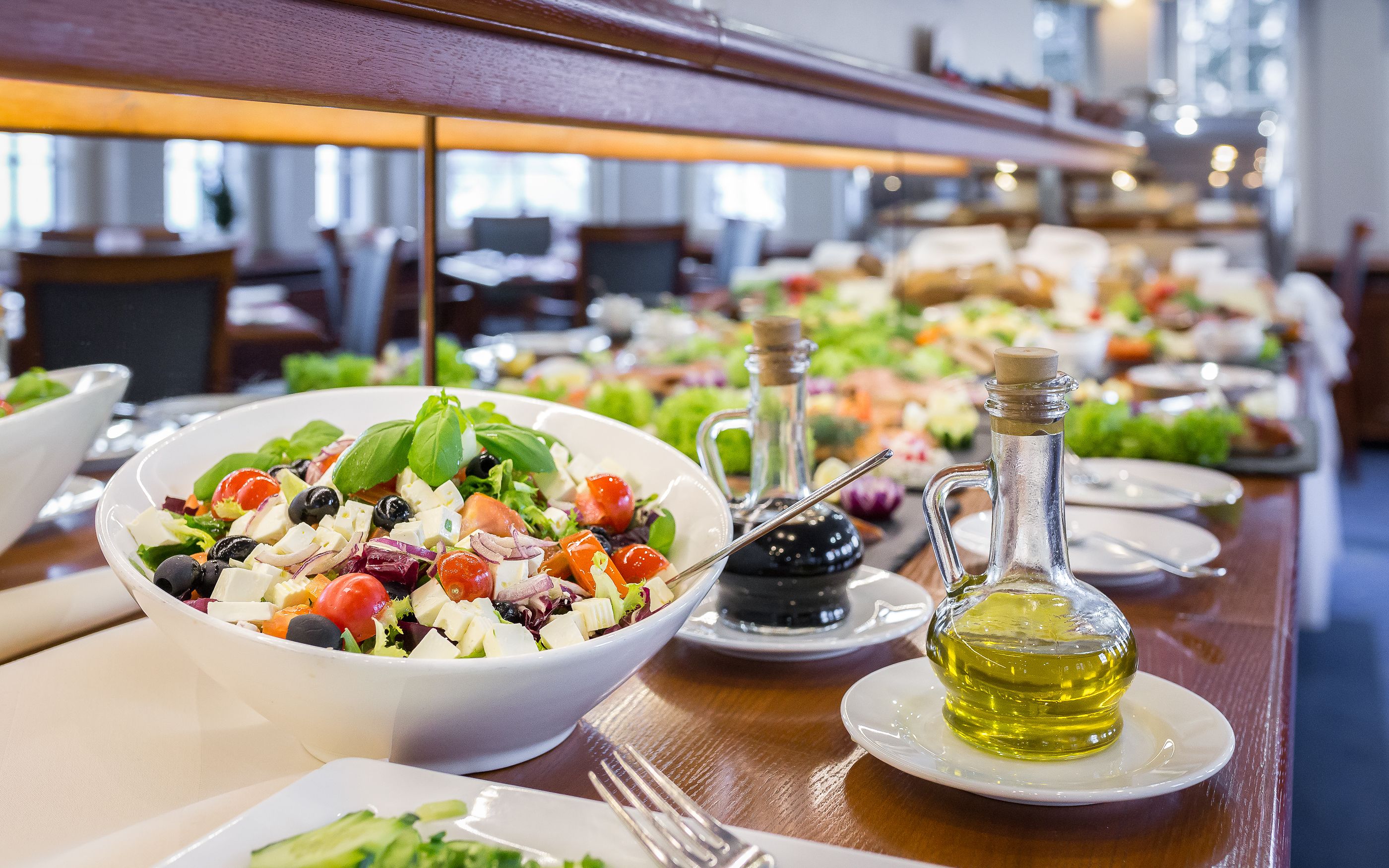 A buffet table with a variety of dishes, including a bowl of salad with tomatoes, olives, and cheese, and a bottle of olive oil.