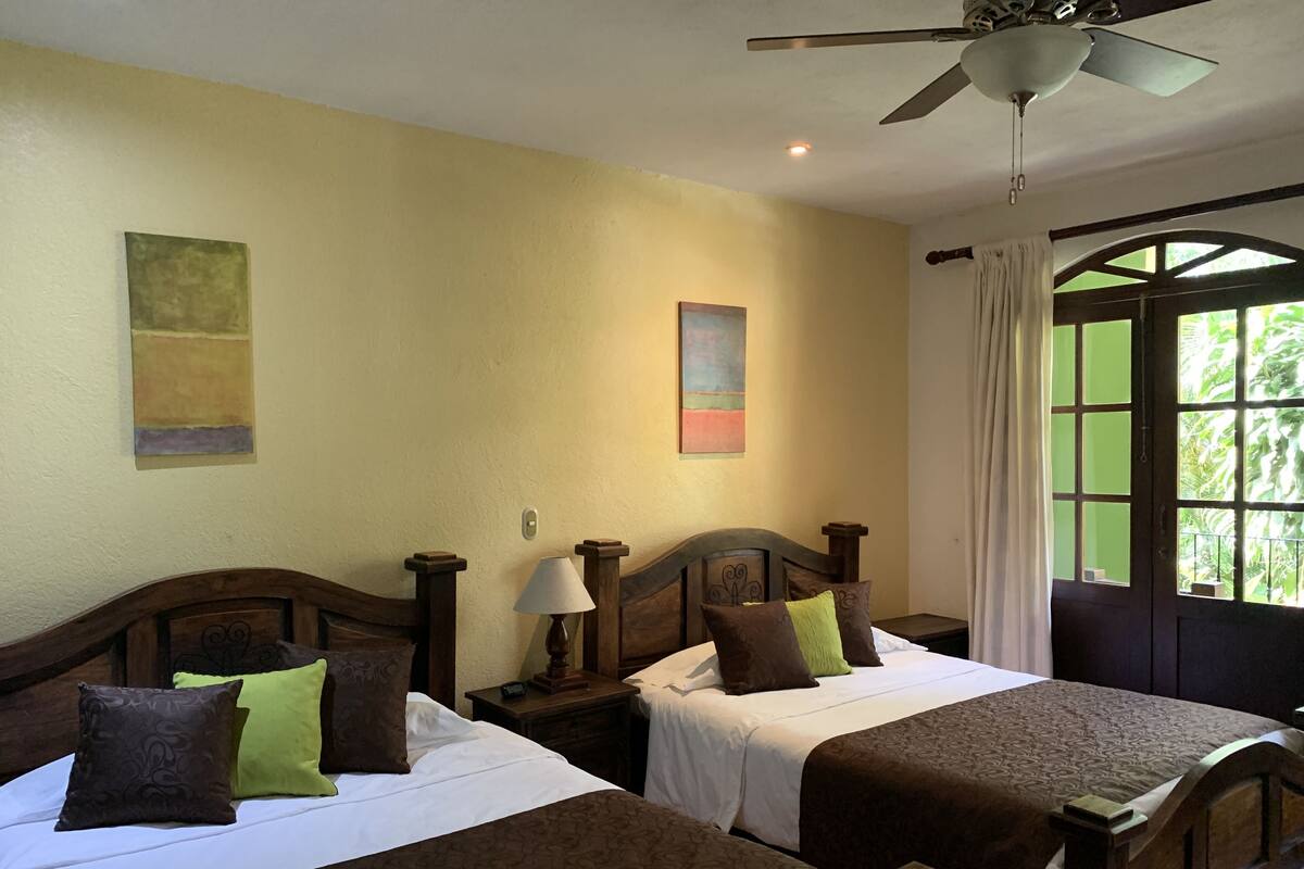 A hotel room with two beds, a ceiling fan, a window with curtains, and artwork on the walls.