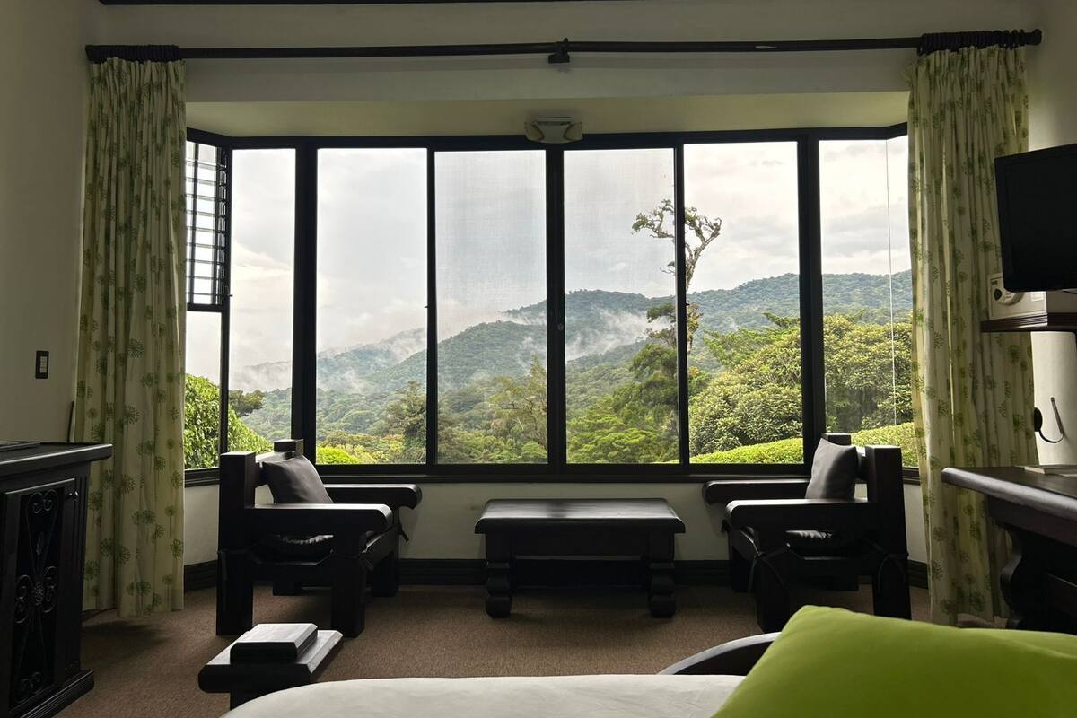 A hotel room with a large window offering a view of a mountainous landscape. There are two black chairs, a bench, and a bed with a green pillow.