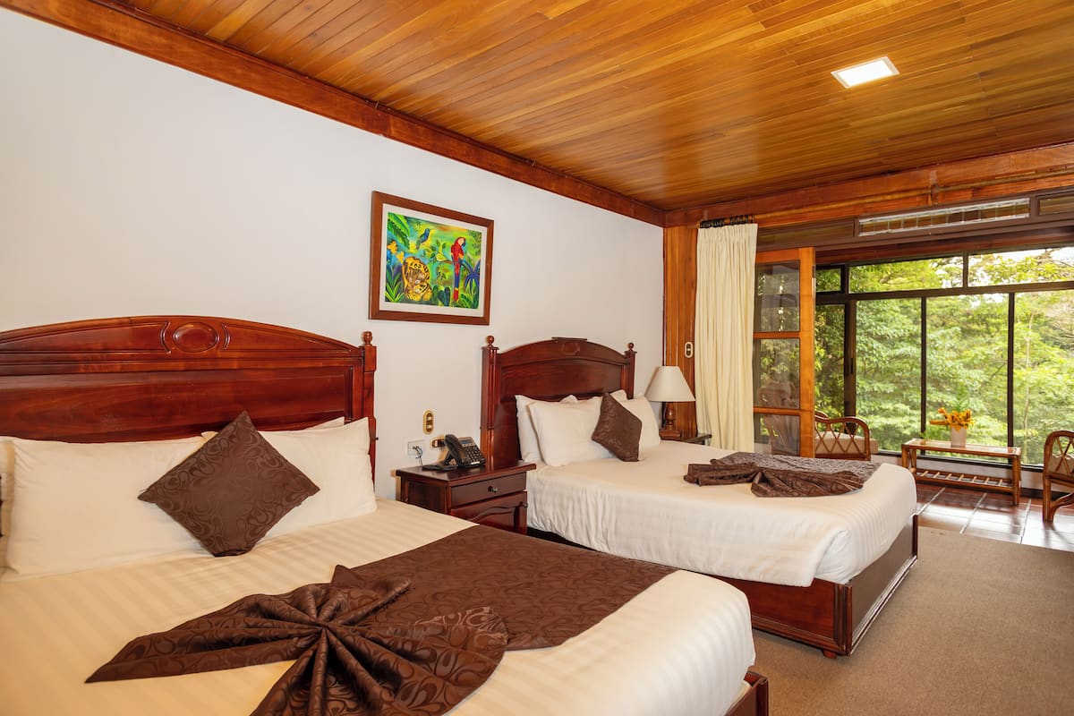 A hotel room with two beds, wooden headboards, a painting on the wall, and a large window with a view of greenery.