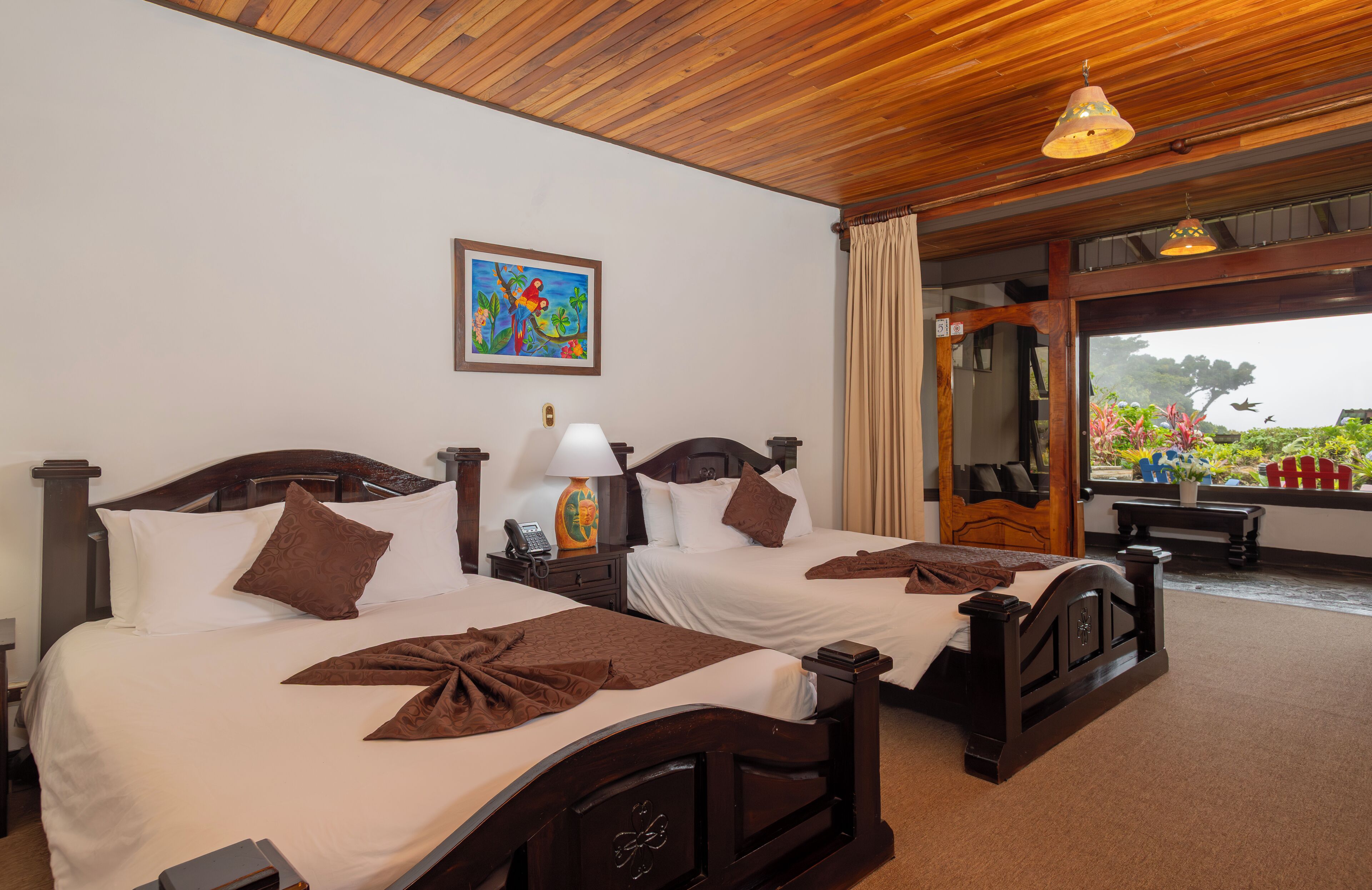 A hotel room with two wooden beds, a painting on the wall, and a view of the outdoors.