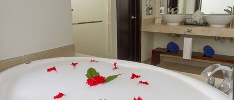 Une salle de bain avec une baignoire remplie d’eau et de pétales de fleurs rouges, un lavabo et un miroir au-dessus.