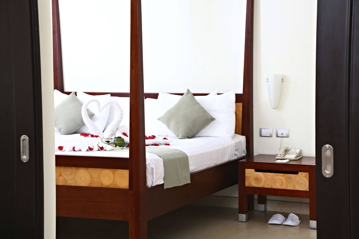 Una cama con dosel, toallas con forma de corazón, una mesita de noche con un teléfono y zapatillas en el suelo.