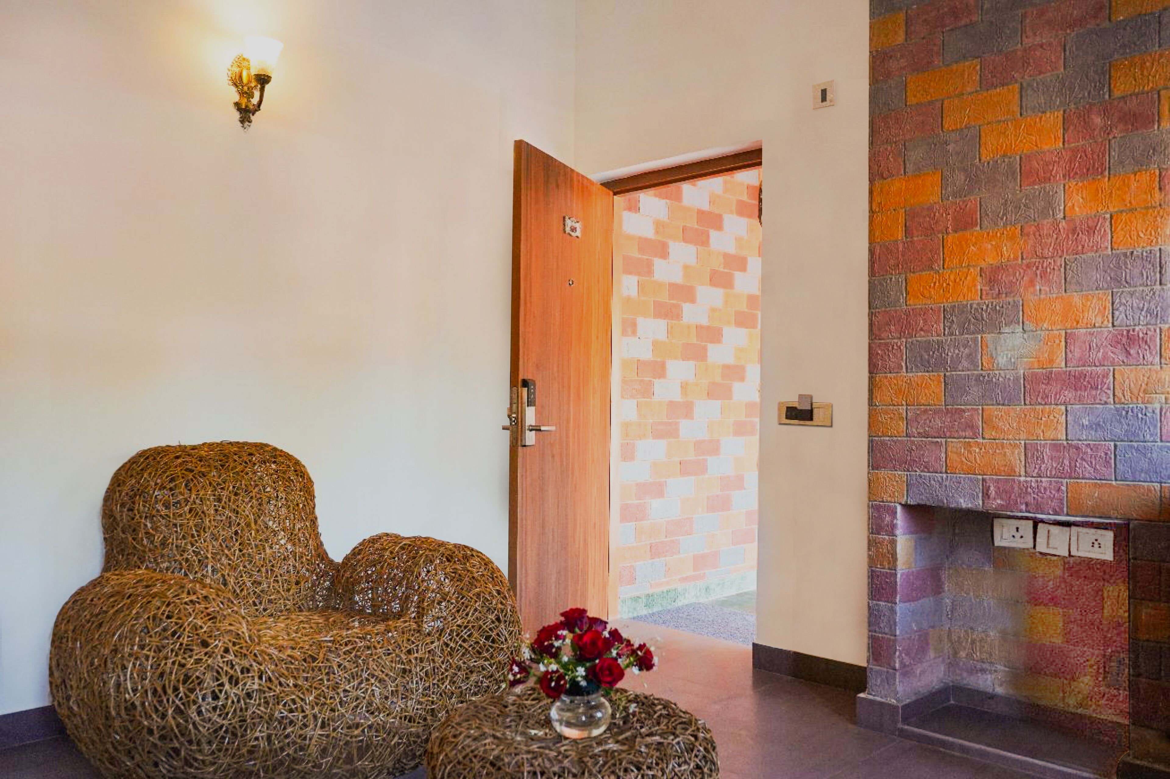 A room with a wicker sofa, a small round table with a vase of flowers, a brick-patterned wall, and a wooden door.
