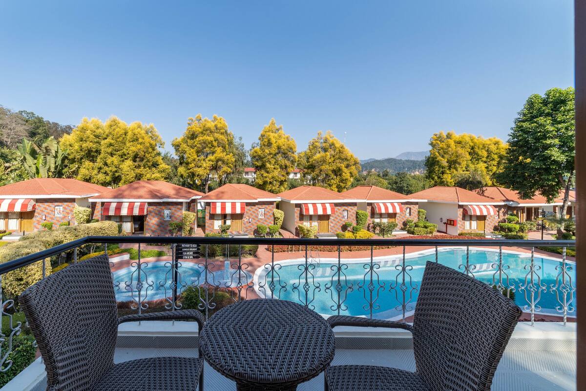 A poolside view with wicker furniture, a clear blue pool, and a row of red-roofed buildings with striped awnings.