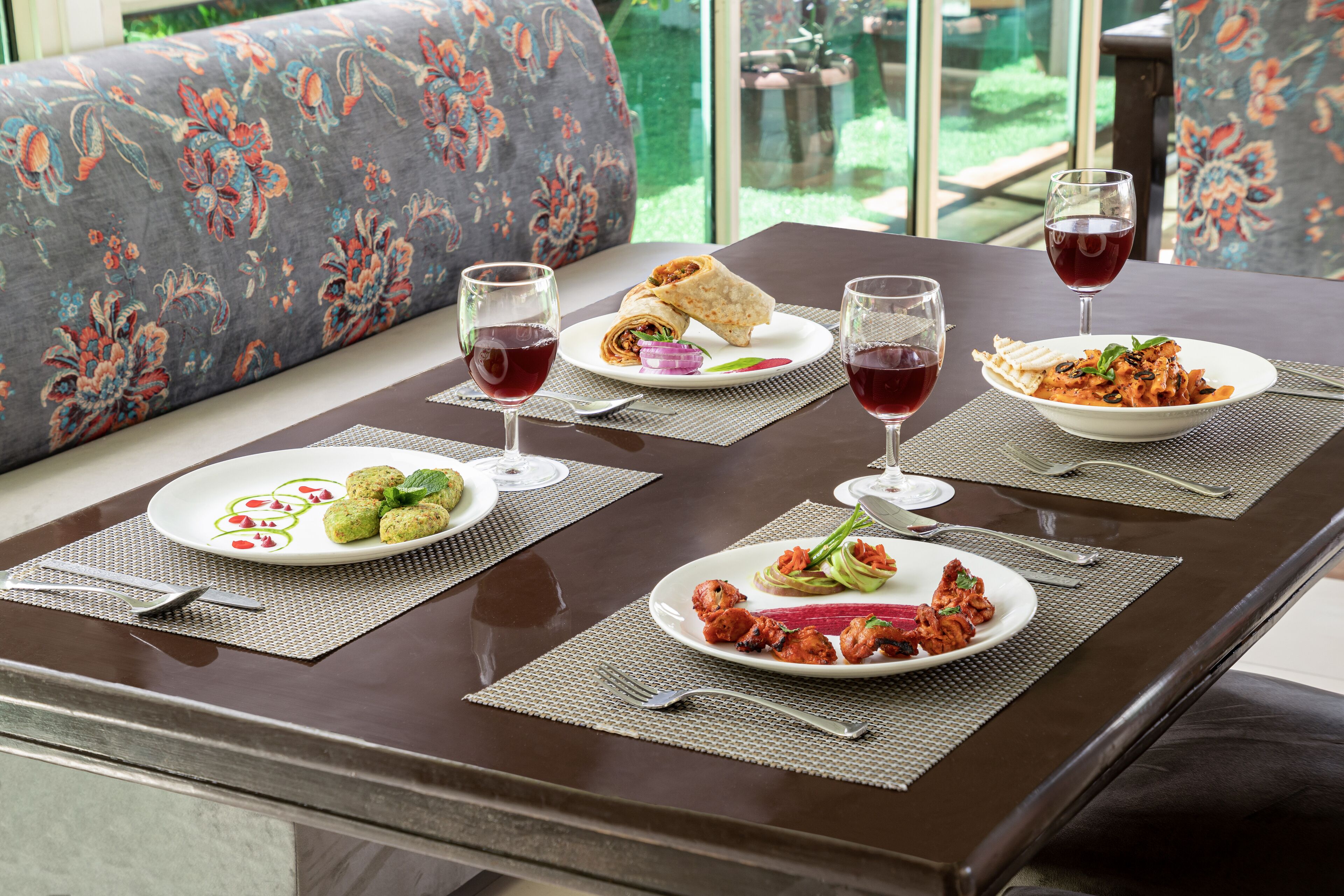 Una mesa de comedor con dos platos de comida, dos copas de vino tinto y un banco con tapicería floral.