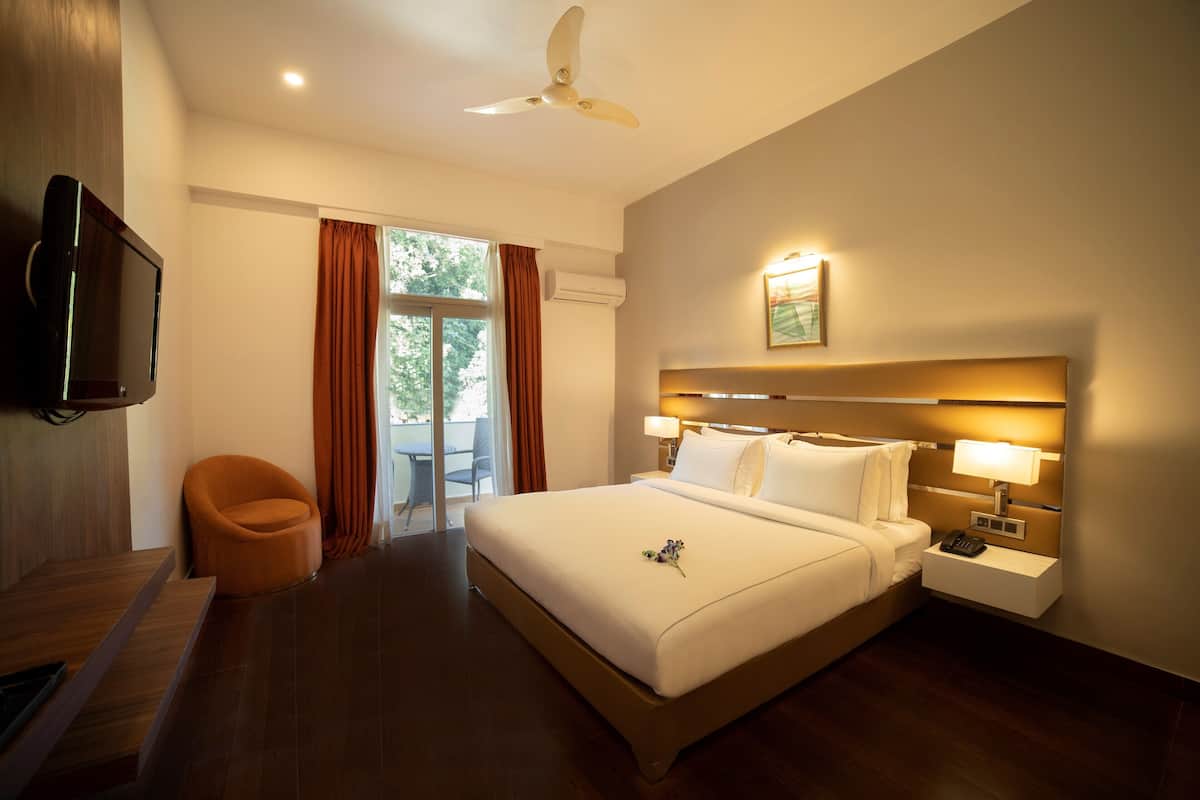 A hotel room with a large bed, two bedside tables with lamps, a flat-screen TV mounted on the wall, a ceiling fan, and a view of the outdoors through a sliding glass door.