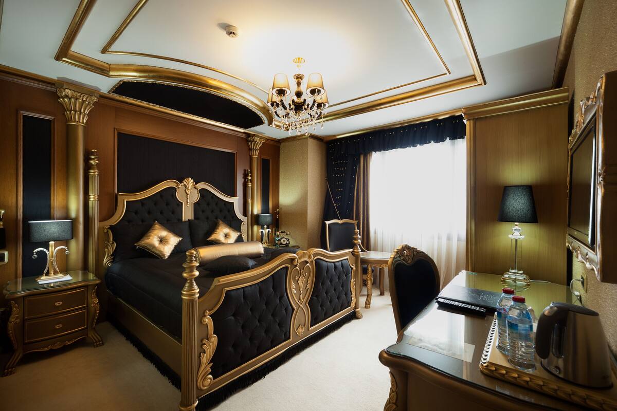A luxurious bedroom with a large bed, a desk, a chair, a chandelier, and a mirror.