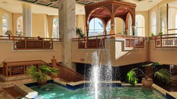 Ein luxuriöser Innenpoolbereich mit einer zentralen Fontäne, einer verzierten Decke und einem Balkon mit Blick auf den Pool.