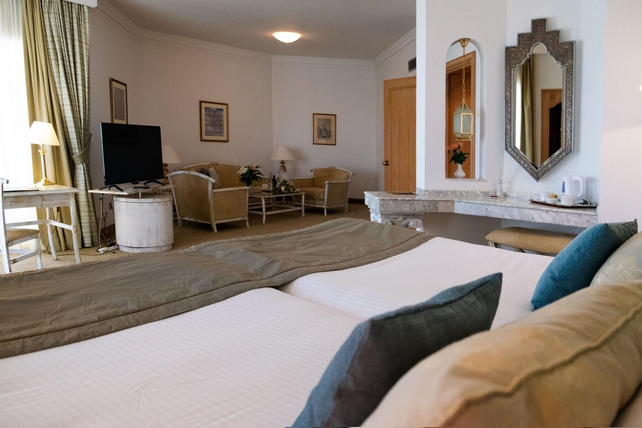 Una habitación de hotel con una cama, dos sillones, un escritorio con televisor, un espejo y una puerta que da a otra habitación.
