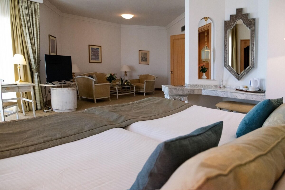 Una habitación de hotel con una cama, dos sillones, un escritorio con televisor, un espejo y una puerta que da a otra habitación.