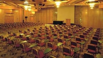 Un amplio salón de conferencias con filas de sillas y mesas, un atril y suelo alfombrado.